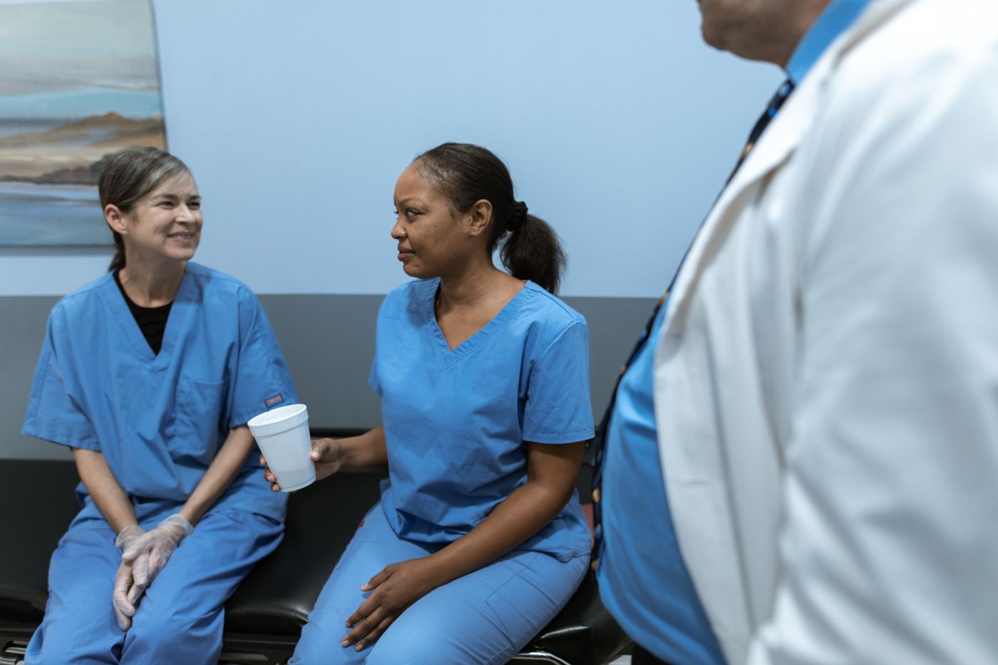 Two nurses in blue scrubs seated and talking with a colleague in a white coat standing beside them in a bright clinical setting
