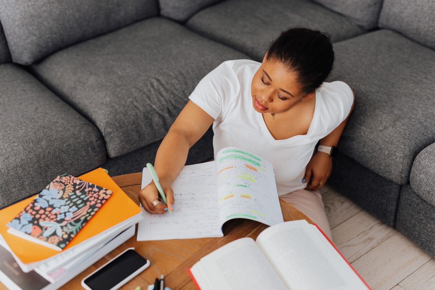 Student sitting on the floor beside a gray sofa writing in a notebook full of highlighted notes, with textbooks, notebooks and a phone on a wooden coffee table
