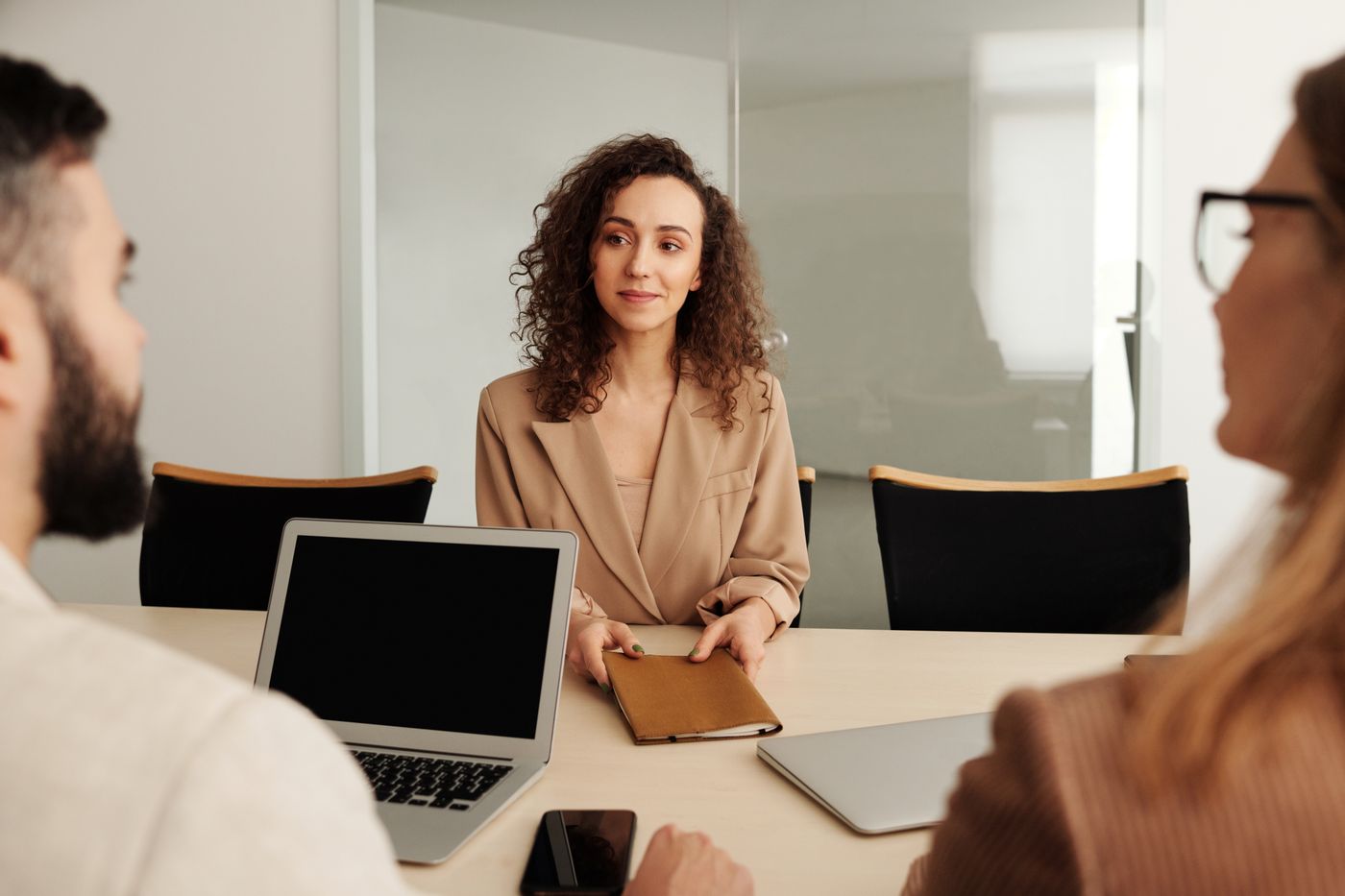 Woman with curly hair in a beige blazer sitting at a conference table in a job interview, holding a folder, facing two interviewers with laptops