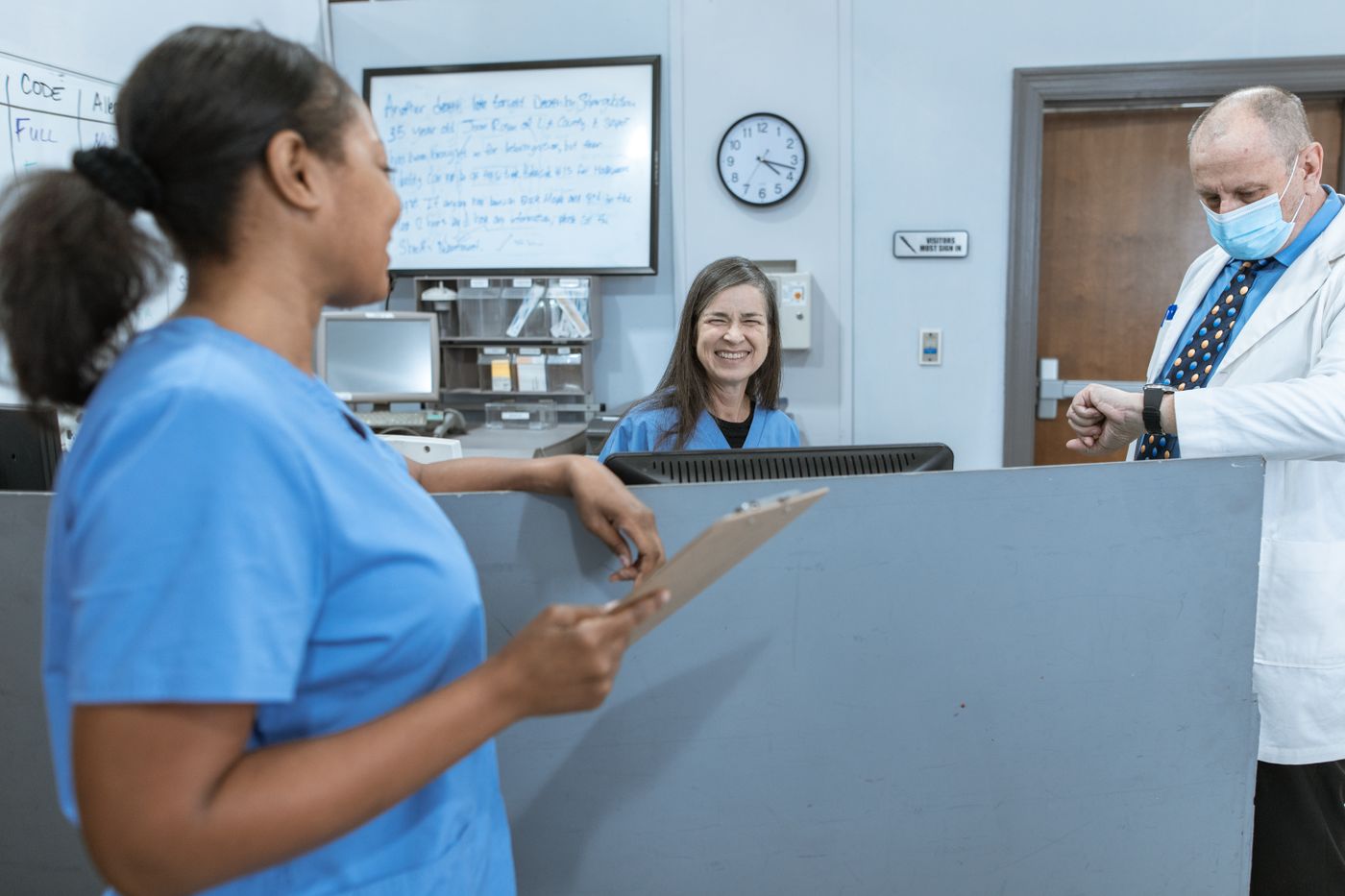 A nurse in blue scrubs holding a clipboard talks with a smiling colleague seated behind the nurses' station, while a physician in a white coat and face mask checks his watch beside a whiteboard