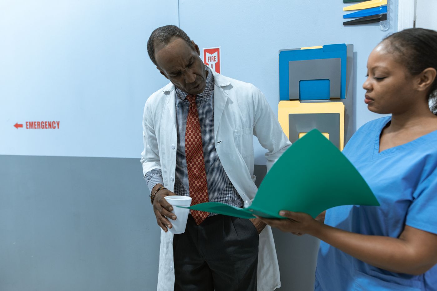 A nurse in blue scrubs holds open a green folder while a senior physician in a white coat and tie leans in to read it, in a hospital corridor with an Emergency sign on the wall