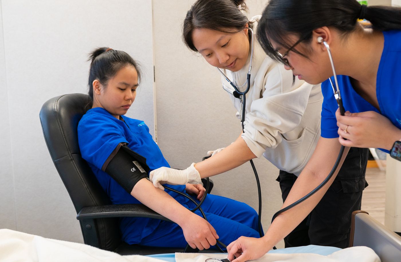 Nursing student in blue scrubs listens with a stethoscope while an instructor guides her hand on a blood pressure cuff on a classmate seated in a chair
