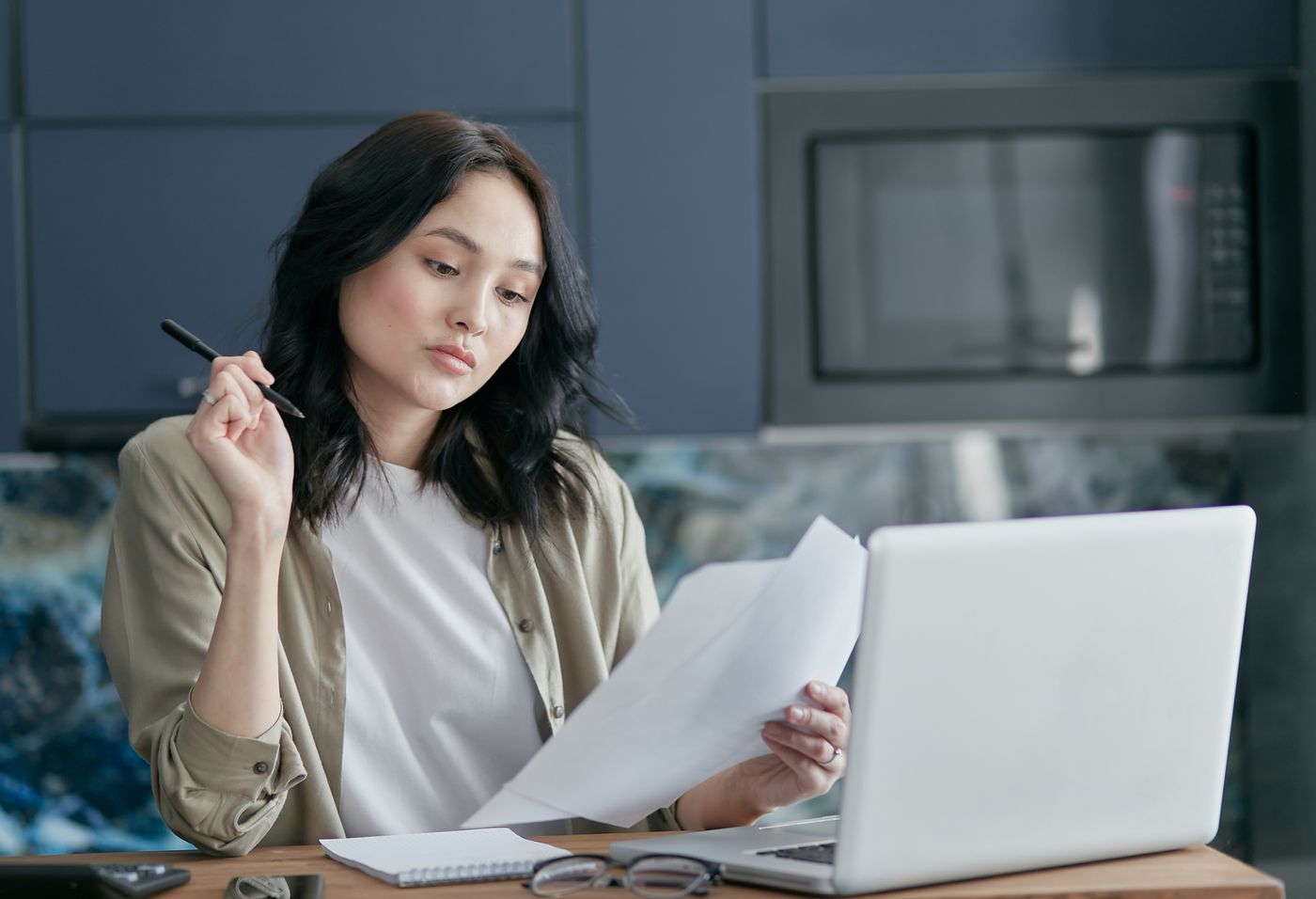 Woman at a kitchen table reading printed papers with a pen in hand, an open laptop and notebook in front of her