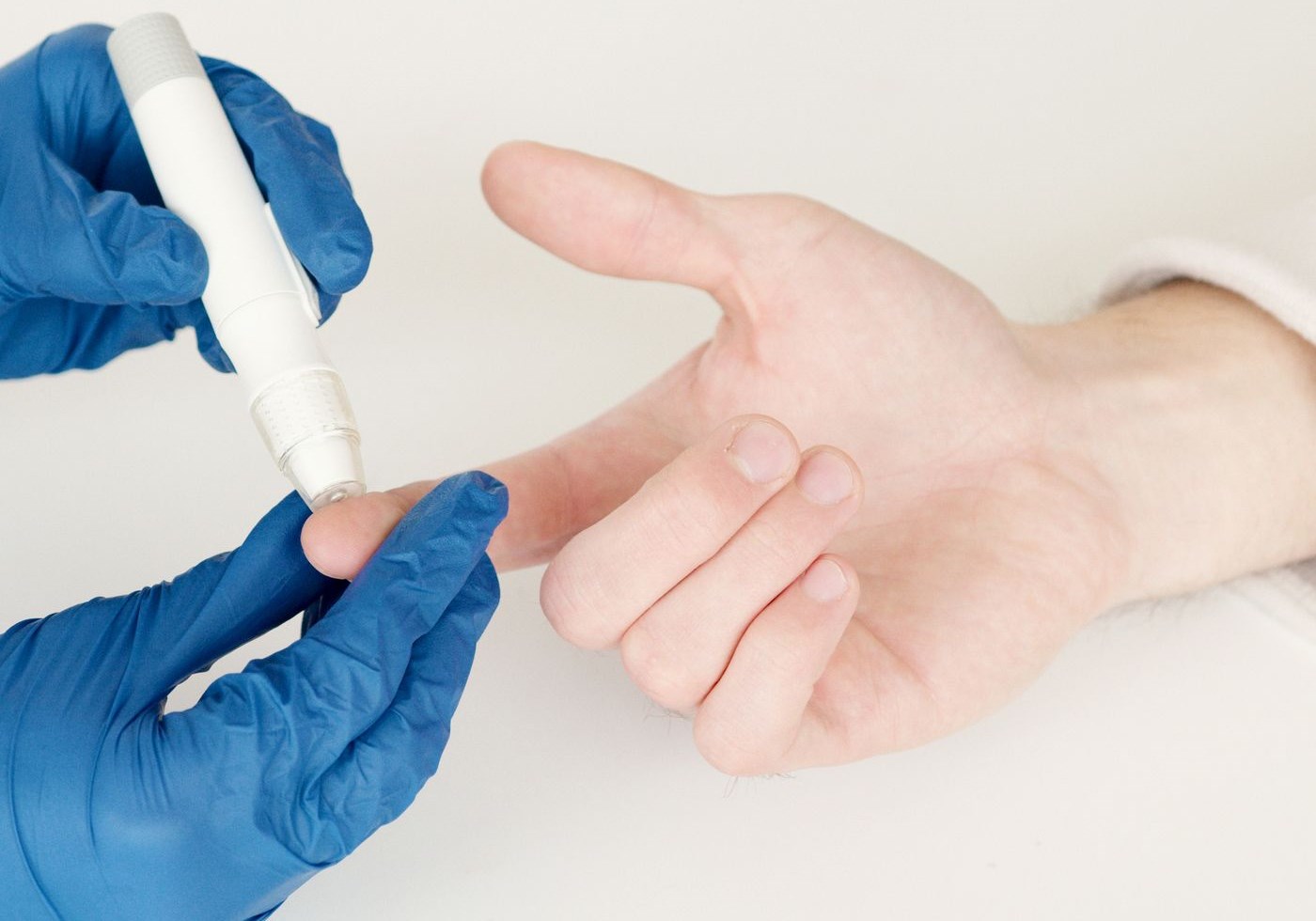Gloved clinician using a lancet pen on a patient's fingertip with a glucose meter resting on the table