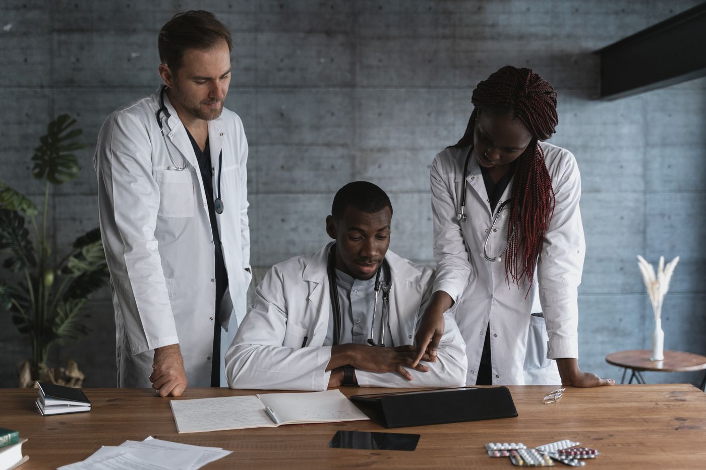 Three clinicians in white coats, a Black woman, a Black man and a white man, reviewing handwritten notes and a tablet together at a wooden table