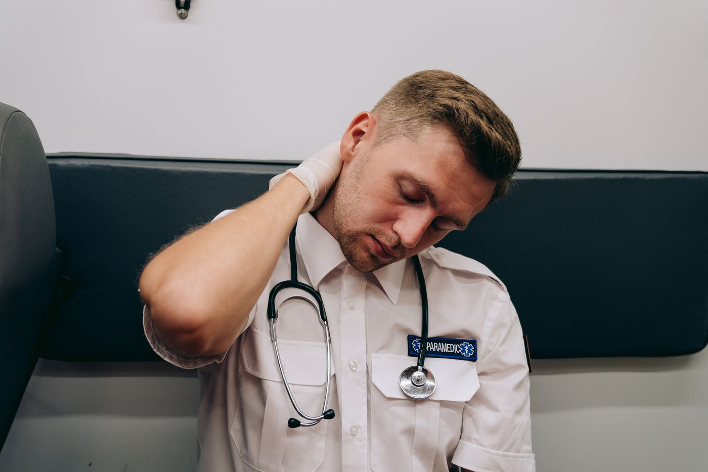 Paramedic in a white uniform shirt with a stethoscope sitting on a stretcher bench, eyes closed, rubbing the back of his neck at the end of a shift