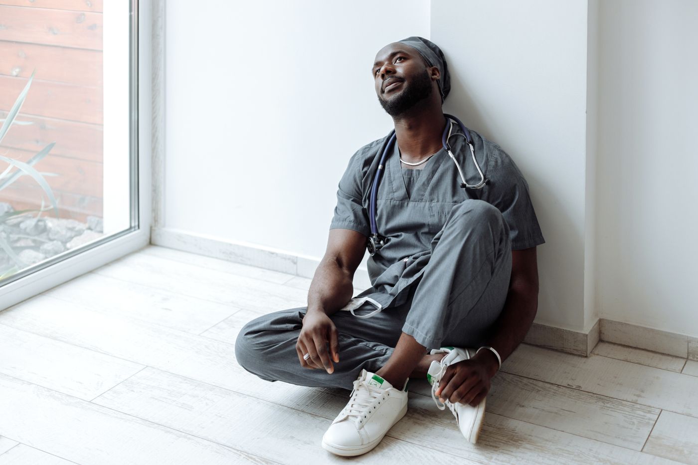 Healthcare worker in gray scrubs with a stethoscope sitting on the floor beside a window, resting against the wall and looking up