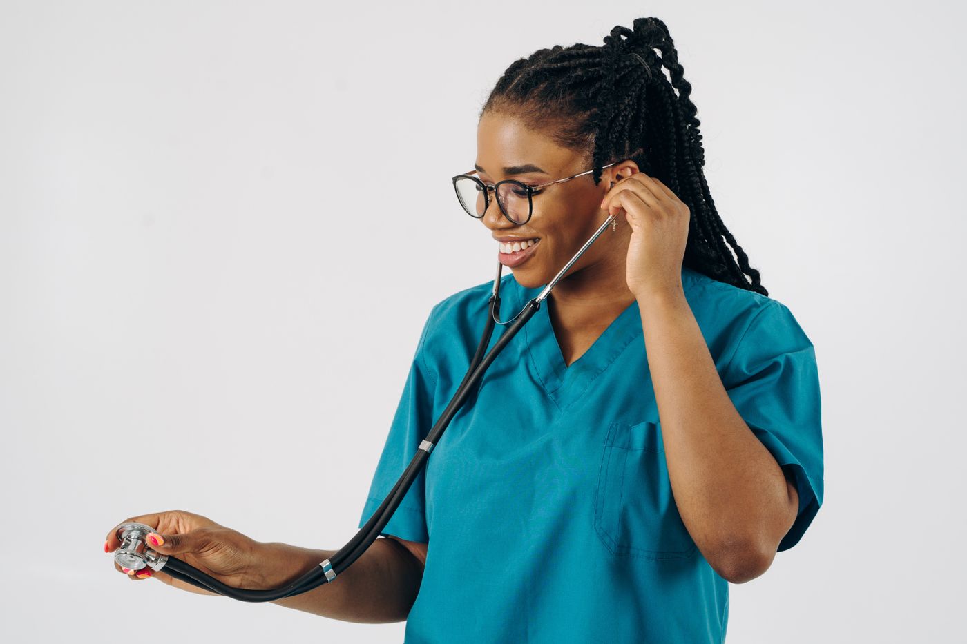 A smiling young nurse in teal scrubs and glasses fitting a stethoscope to her ears against a plain light background