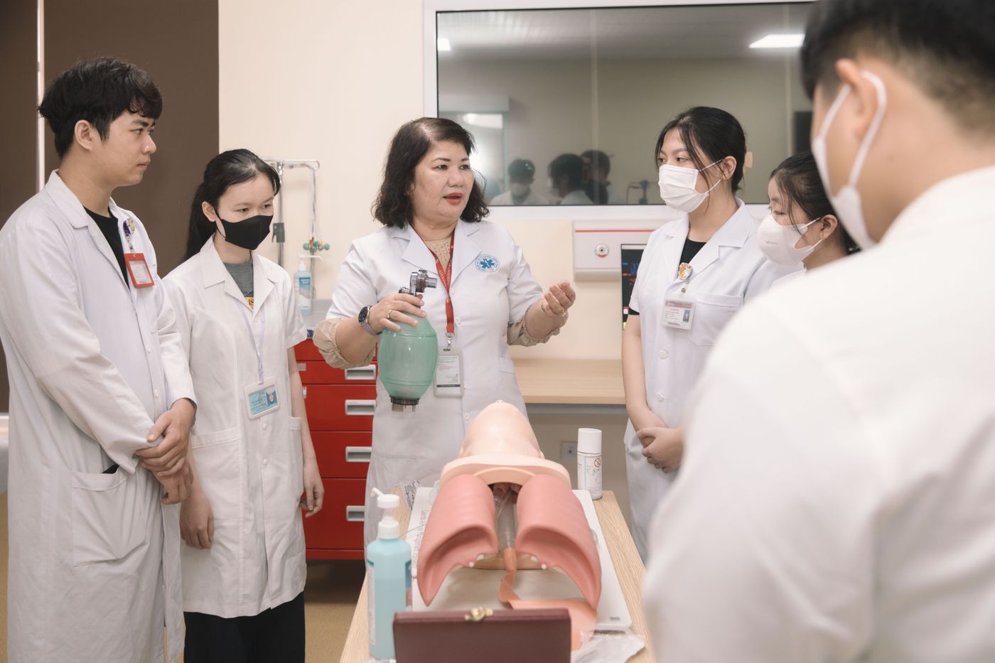 A clinical educator holding a bag-valve resuscitation mask demonstrates airway management to a small group of healthcare staff gathered around an airway training manikin in a facility skills room