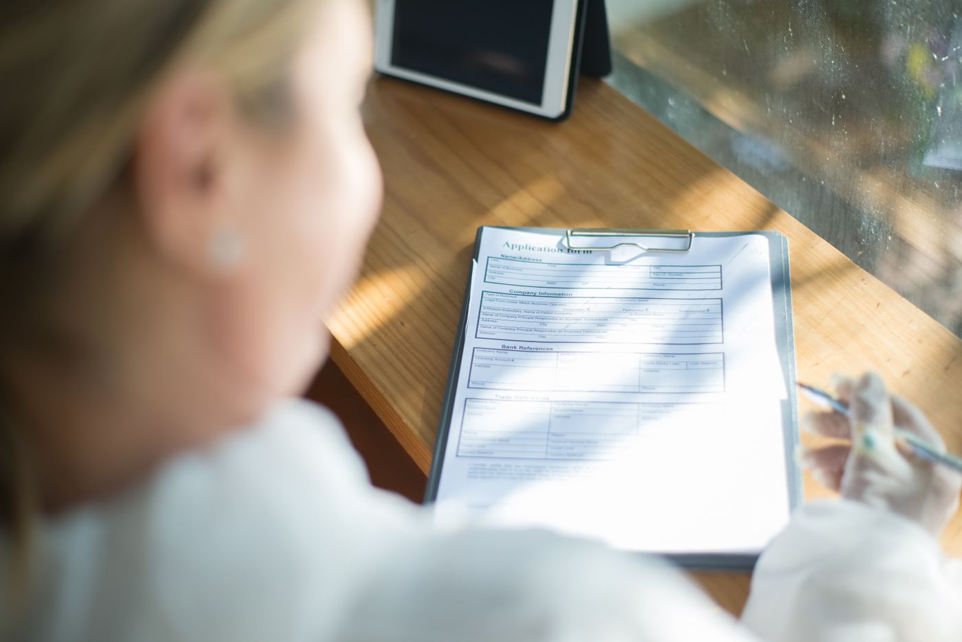 Close-up of a blank application form on a clipboard on a sunlit wooden desk, with a person holding a pen beside it and a tablet in the background