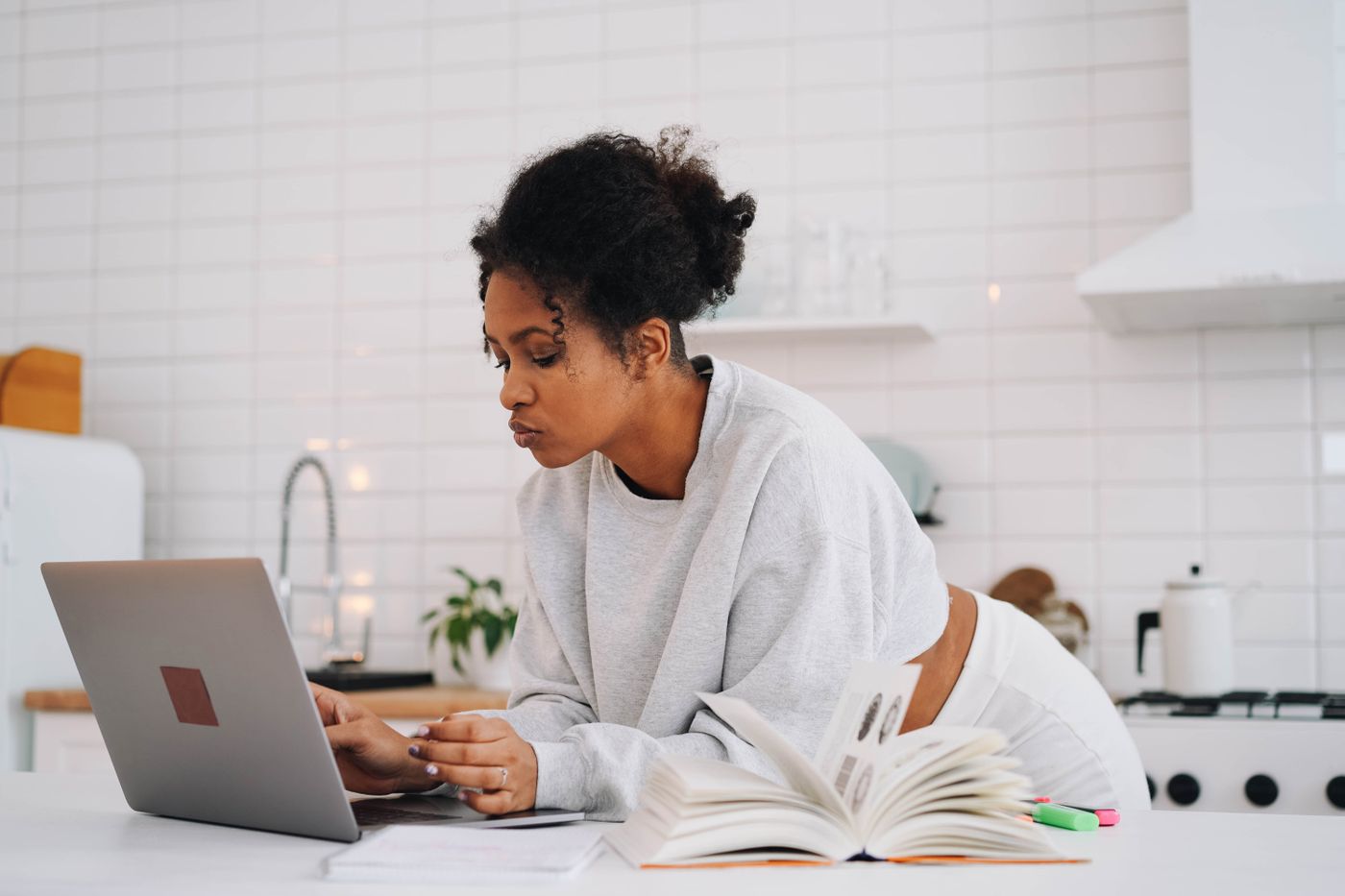 Woman in a gray sweatshirt leans over a white kitchen counter, studying on a laptop beside an open book and highlighters in a bright home kitchen