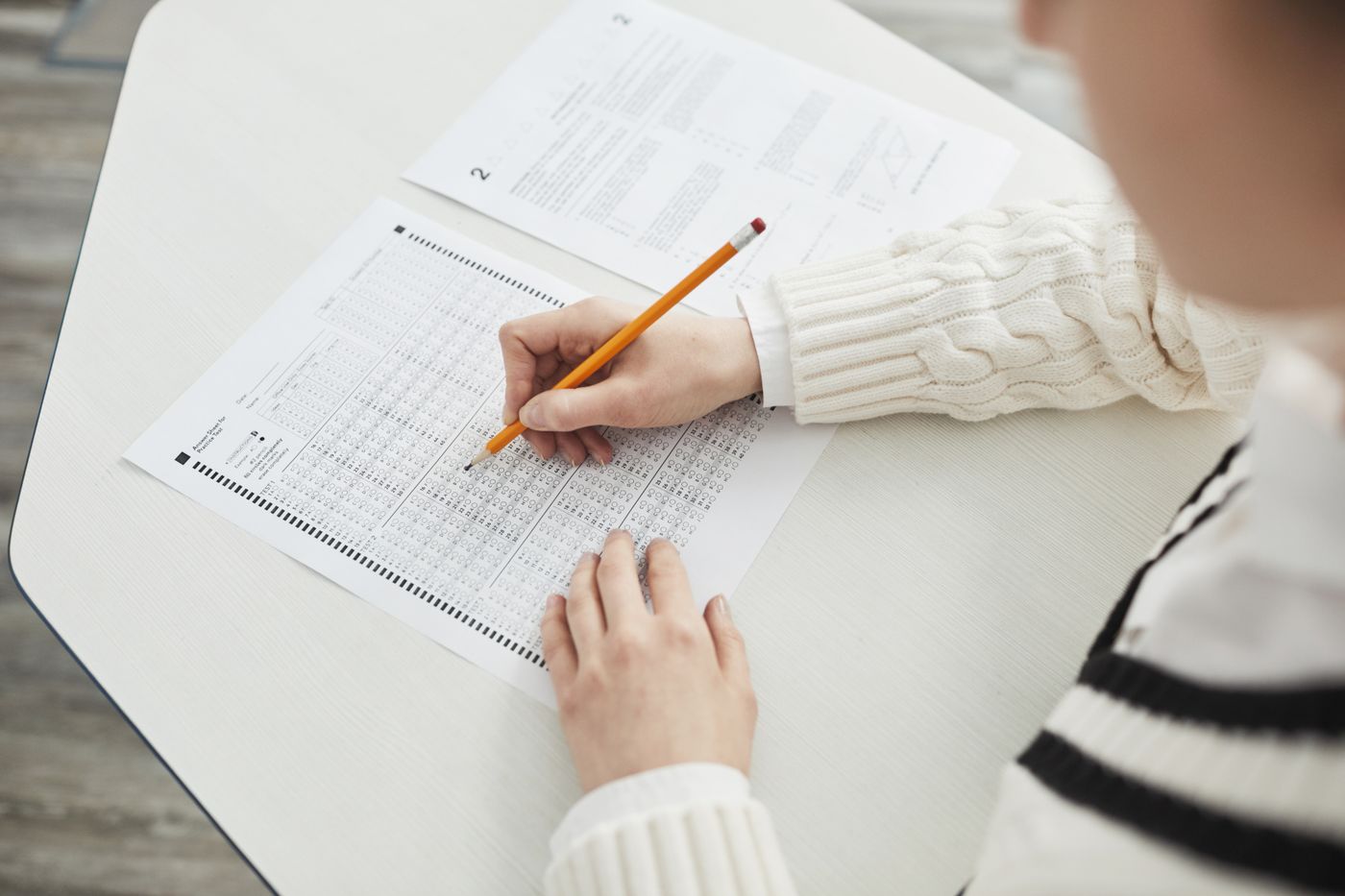 A student in a cream sweater filling in a multiple-choice answer sheet with an orange pencil at a pale desk, a second question paper beside it