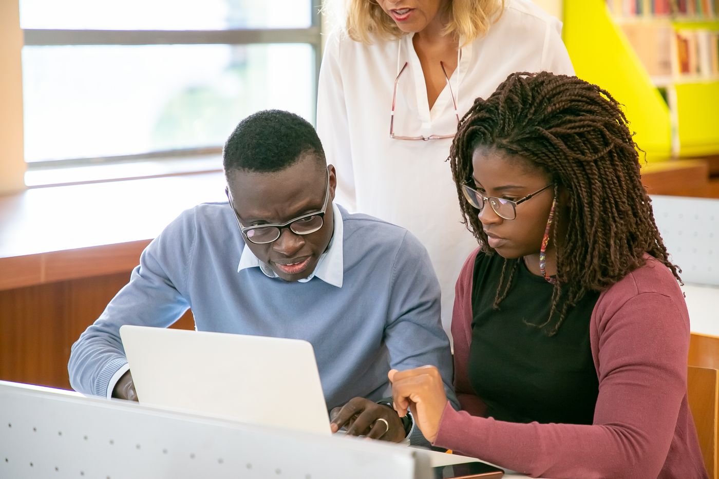 Two Black students in glasses working through a question on a laptop together while an instructor looks on