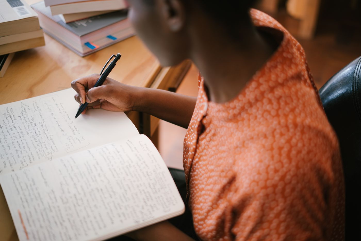 A Black woman writing detailed study notes in an open notebook beside a stack of textbooks