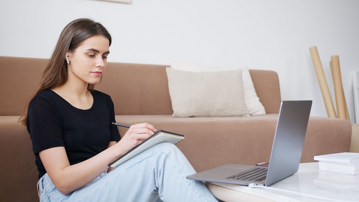 Young woman sitting on the floor by a beige sofa, writing in a notebook beside an open laptop on a coffee table at home