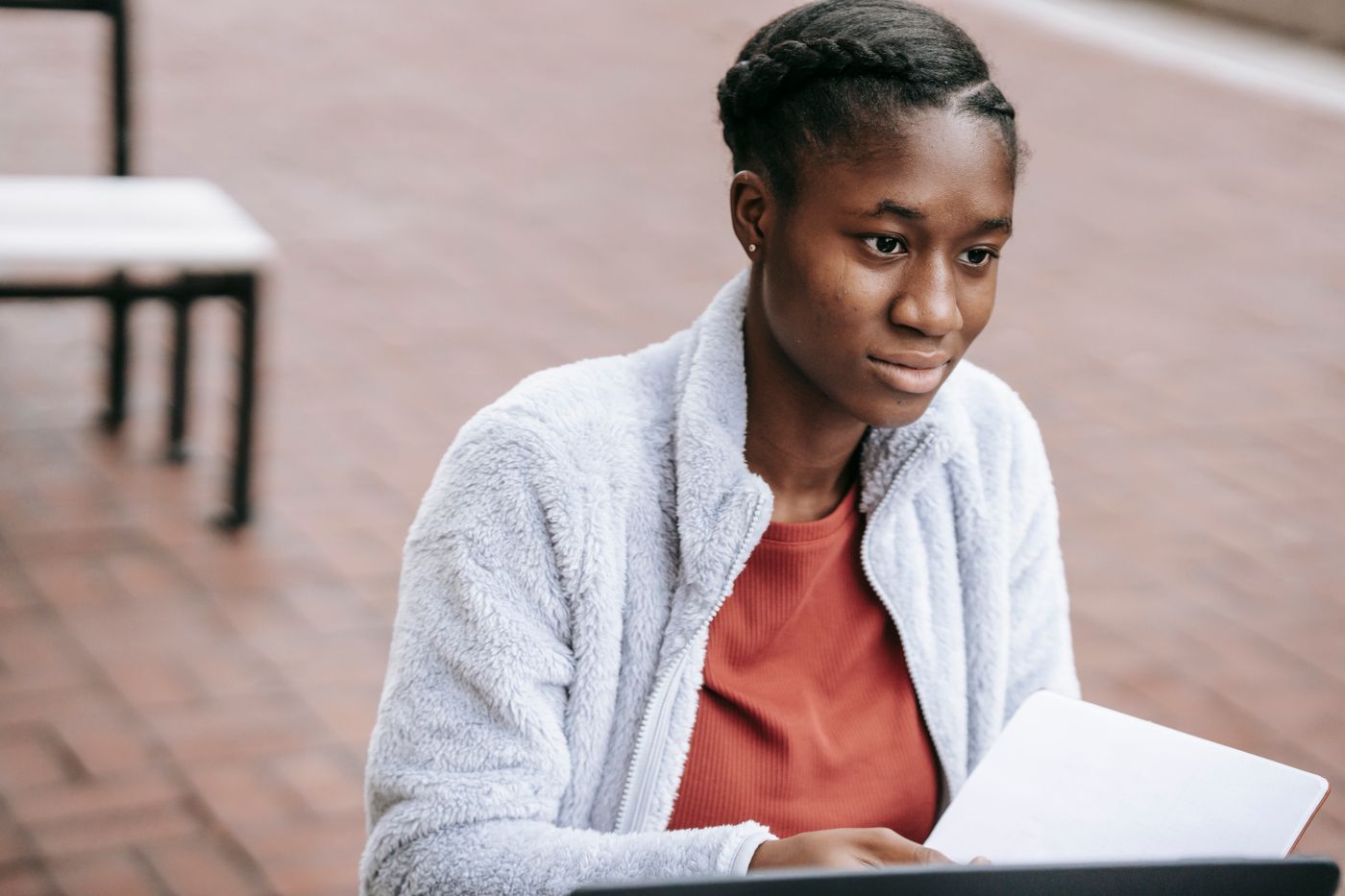 Young Black woman with braided hair in a fleece jacket sitting outdoors, looking thoughtfully at a laptop screen with a notebook beside her