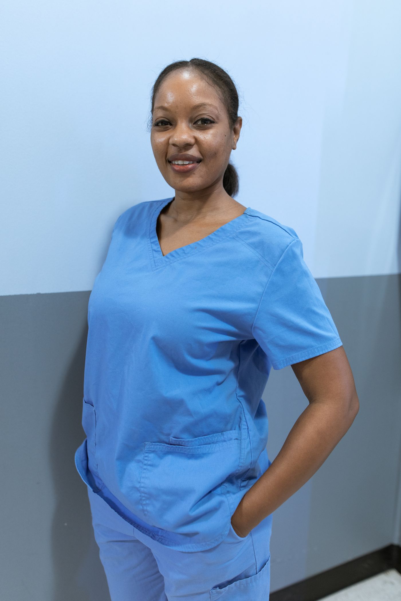 A Black registered nurse in blue scrubs standing confidently against a clinic corridor wall
