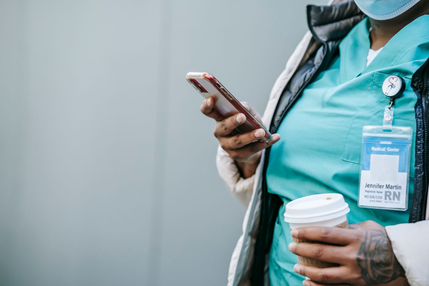 Close-up of a nurse in teal scrubs and a winter jacket holding a smartphone in one hand and a takeaway coffee cup in the other, with a hospital ID badge marked RN clipped to the scrub top