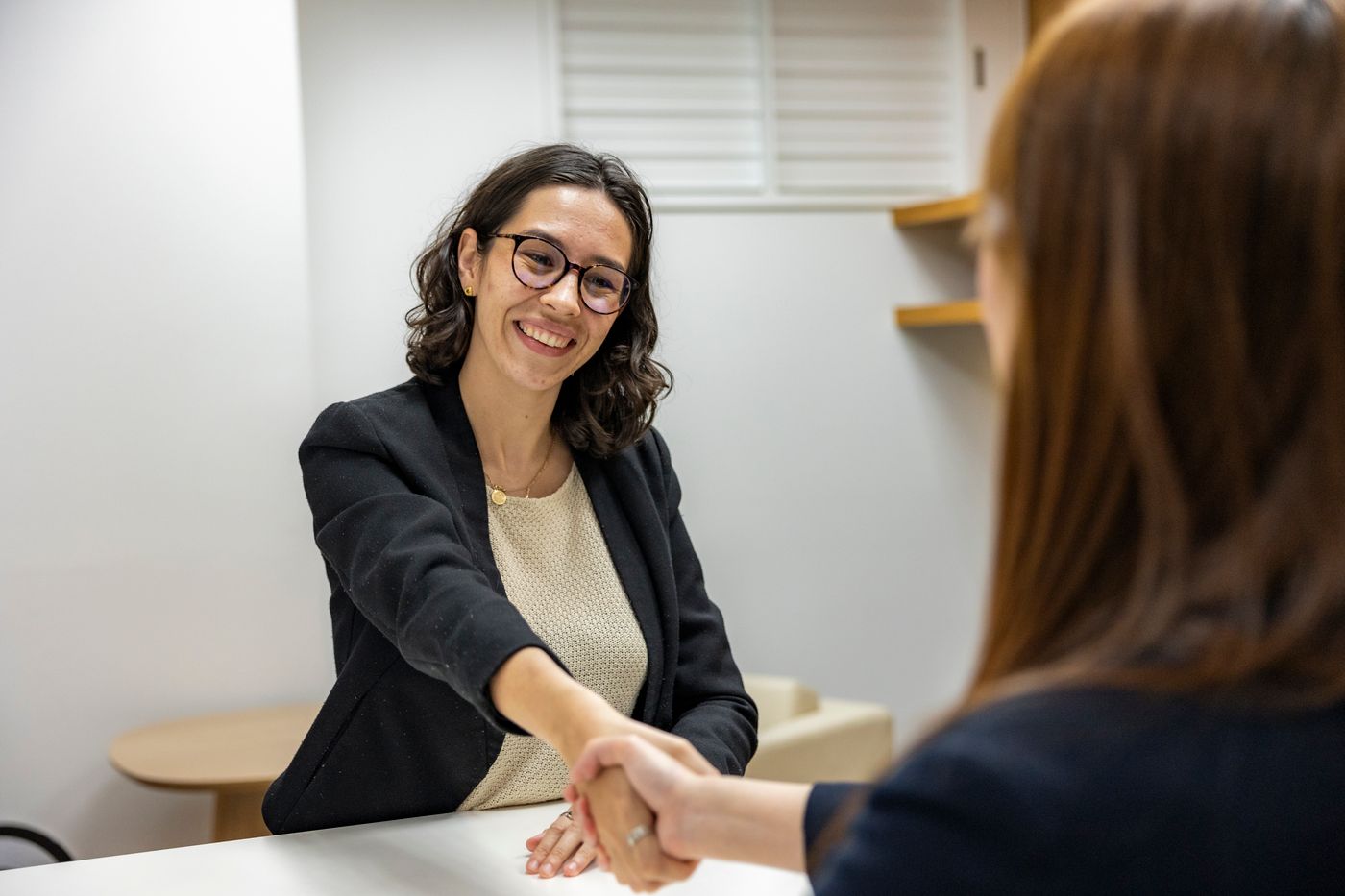 Smiling young woman in glasses and a dark blazer shaking hands across a white table with an interviewer seen from behind, in a bright office