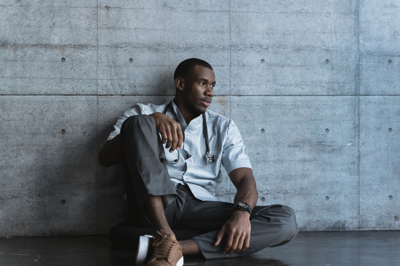 Young clinician in scrubs with a stethoscope sitting on the floor against a concrete wall, looking away in thought