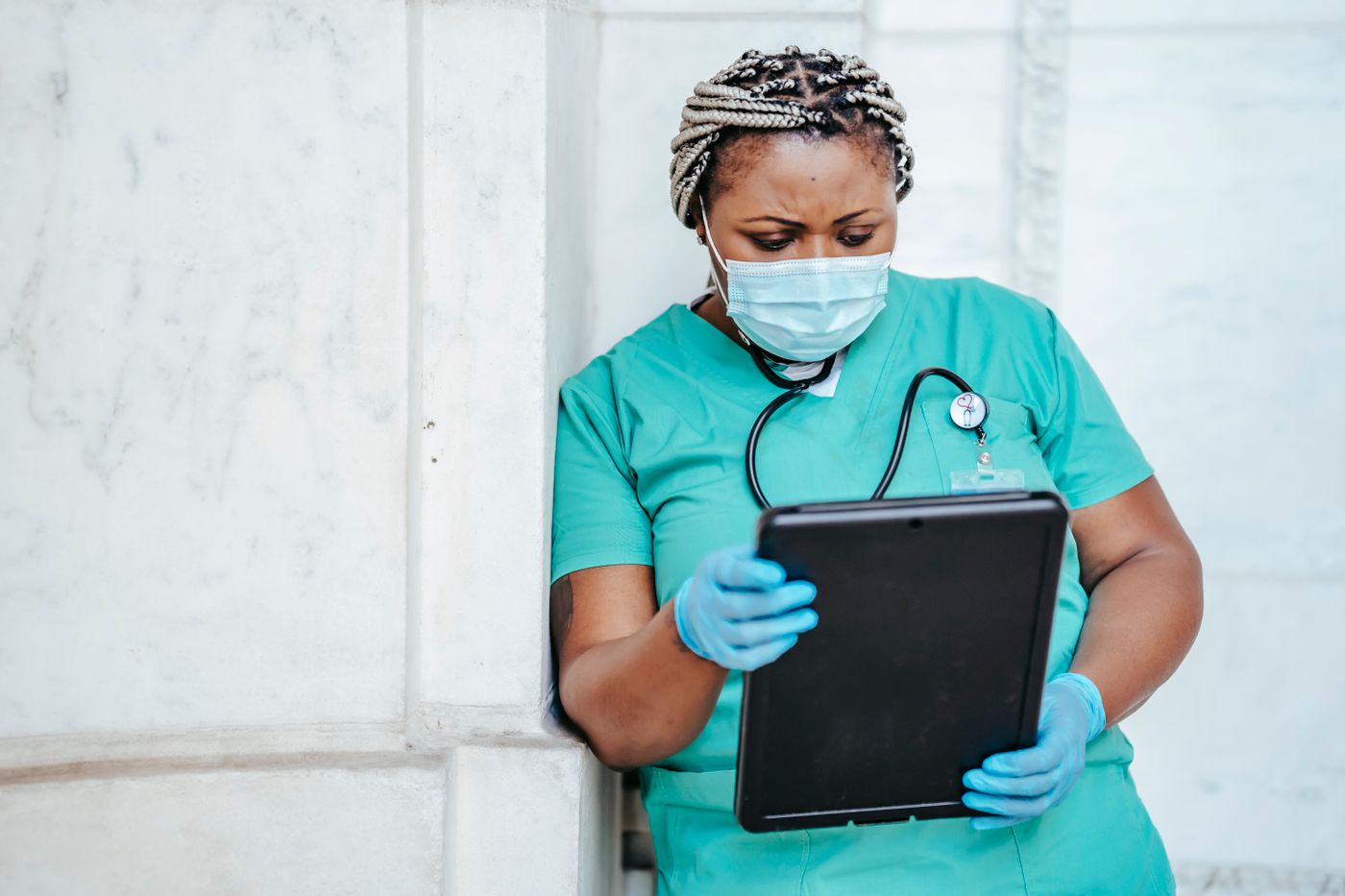 Nurse in green scrubs and a face mask leaning against a wall, reading a tablet with a concerned expression
