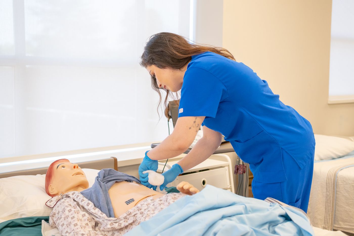 A nurse in blue scrubs and gloves working at the abdomen of an adult patient simulation manikin lying in a hospital bed in a bright skills lab