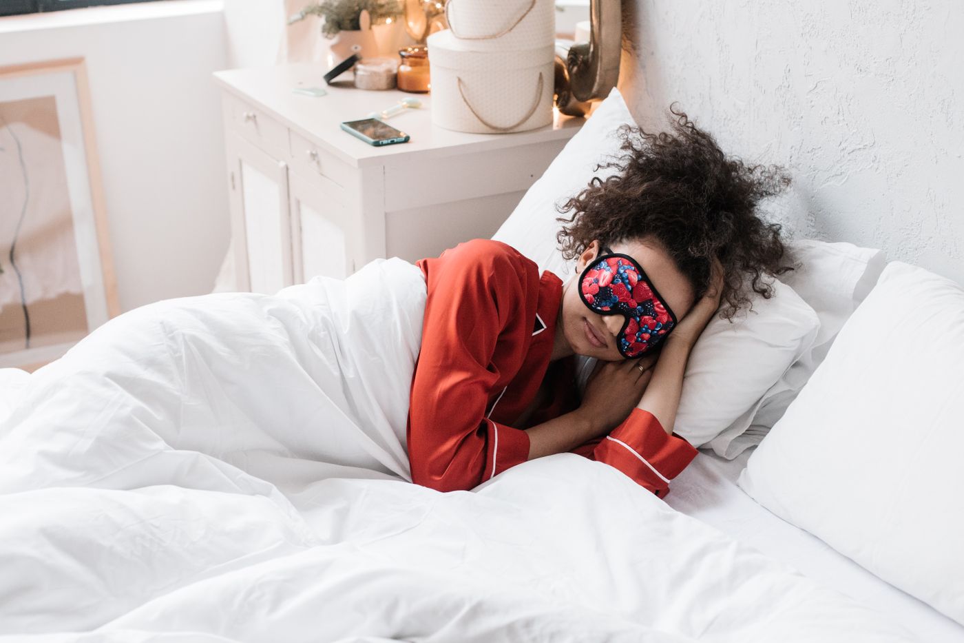Woman with curly hair, wearing red pajamas and a patterned sleep mask, asleep under a white duvet in a bright bedroom during the day