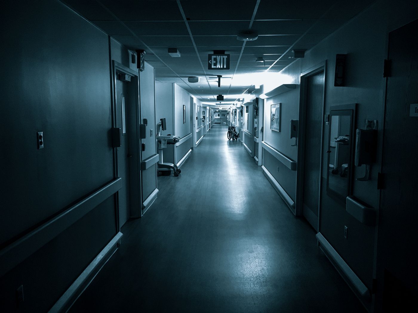 Darkened hospital ward corridor at night, lit by a single bank of ceiling lights and a glowing exit sign, with patient room doors, handrails and an empty wheelchair further down the hall