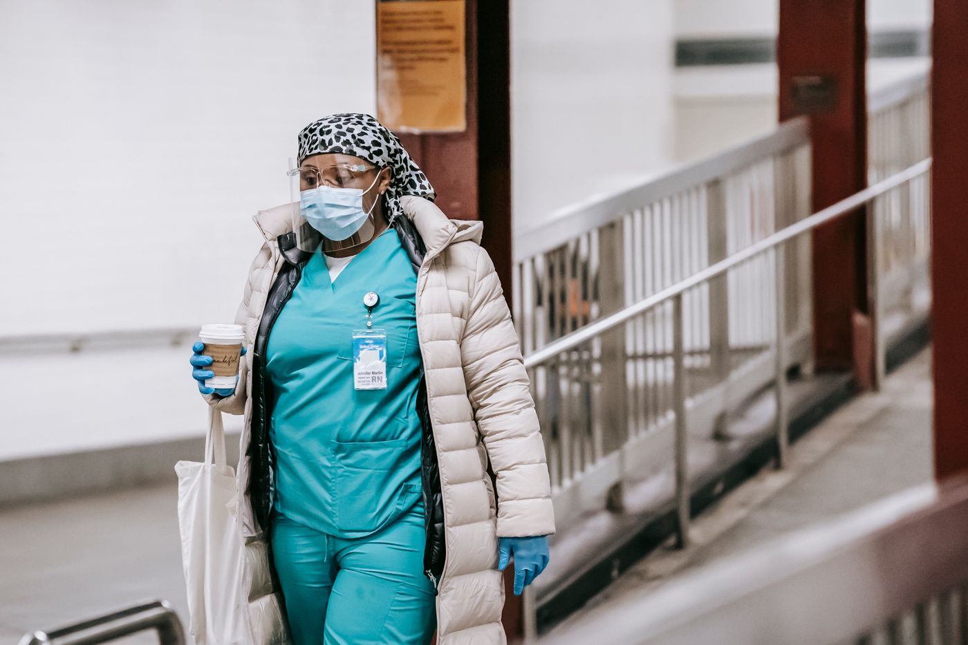 Nurse in teal scrubs and a winter coat walking through a transit station with a coffee cup and a tote bag on the way to or from a shift