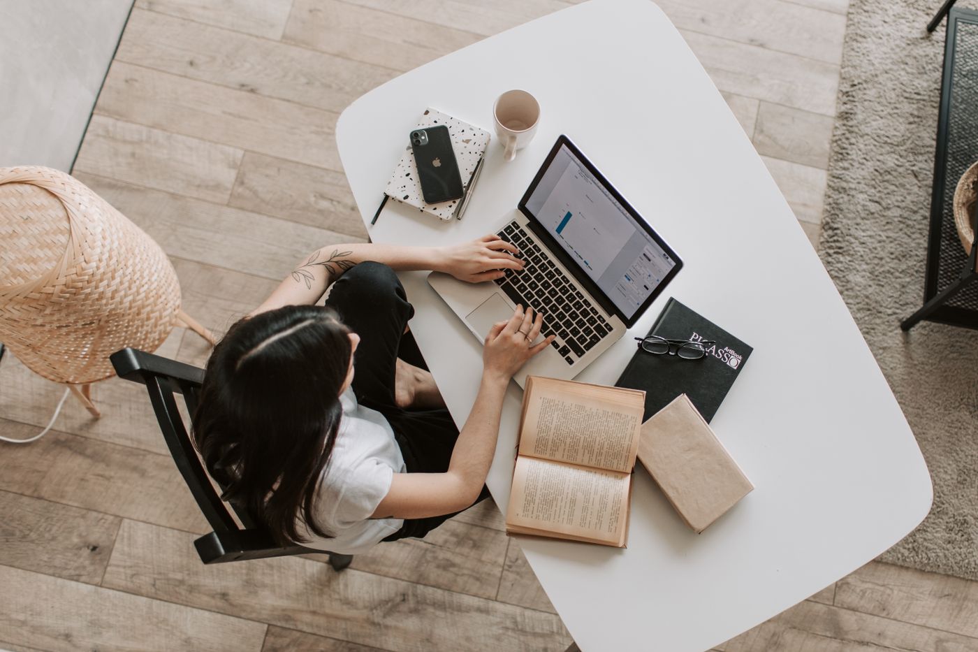 Overhead view of a young woman with long dark hair typing on a laptop at a white table at home, with a phone, a notebook, a coffee mug, glasses and two open books beside her