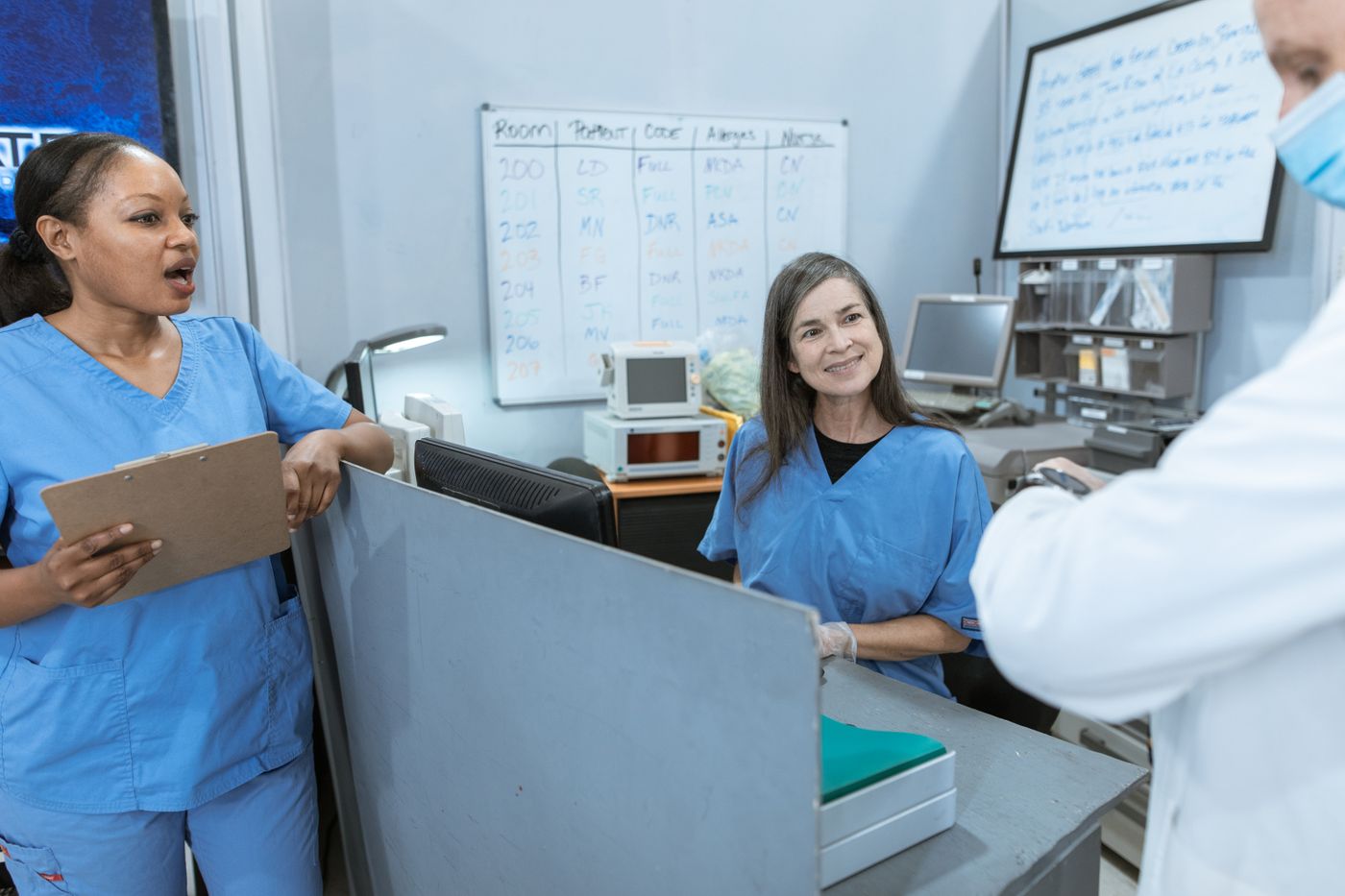 Two nurses in blue scrubs at a hospital nurses station with a patient assignment whiteboard behind them, one speaking while the other listens