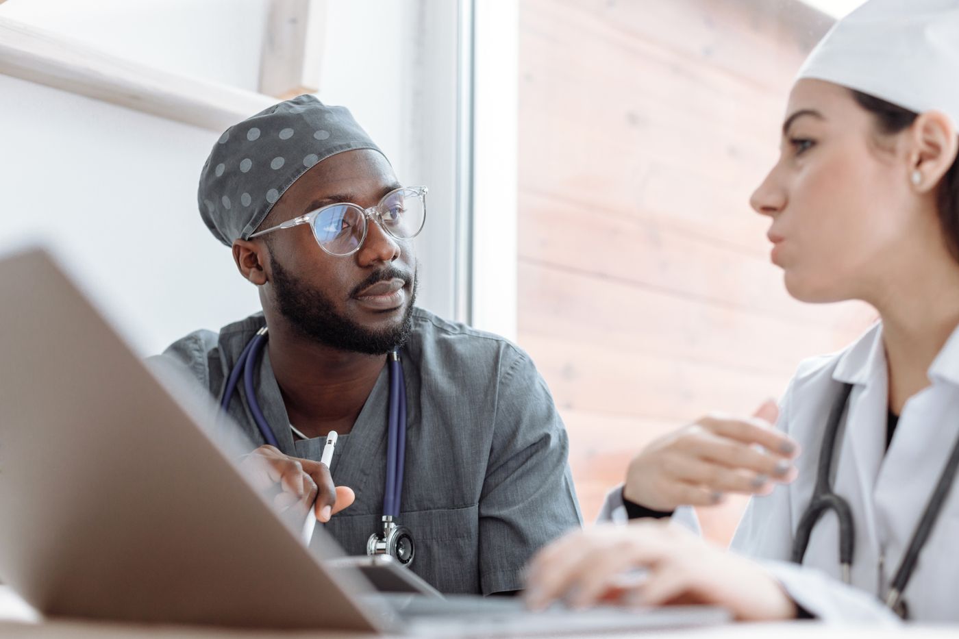Two clinicians in conversation at a desk: a Black man in gray scrubs, a patterned scrub cap and glasses listens closely while a colleague in a white coat and cap explains something beside an open laptop
