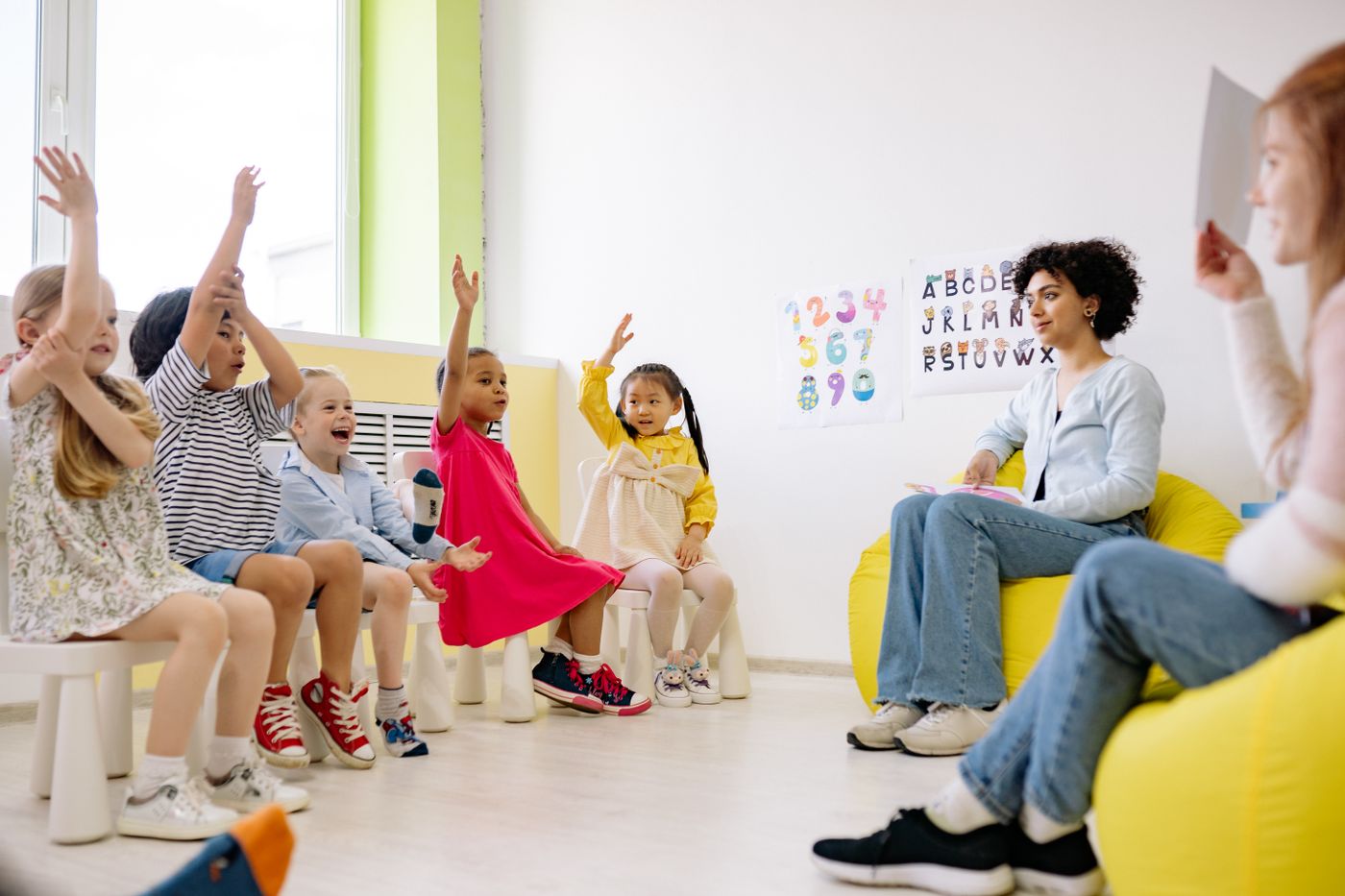 Two caregivers sitting on a yellow beanbag lead circle time in a bright child care classroom while a diverse group of young children on small white chairs raise their hands