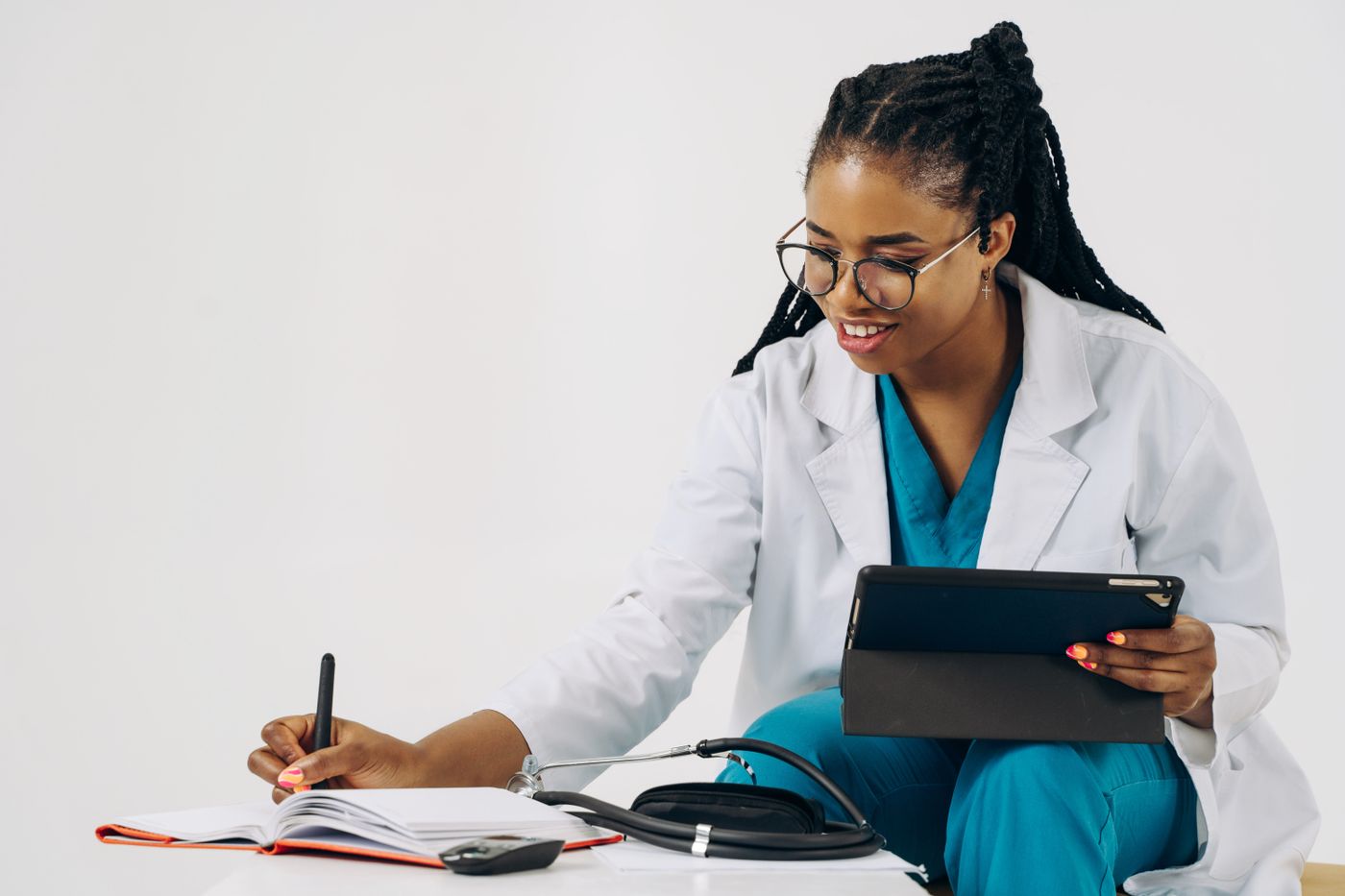 A smiling Black healthcare professional in teal scrubs and a white coat writes in a notebook while holding a tablet, with a stethoscope and a phone on the desk beside her