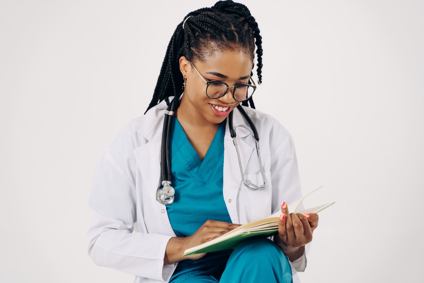 A smiling clinician in teal scrubs and a white coat, with a stethoscope around her neck, reading an open notebook against a plain white background
