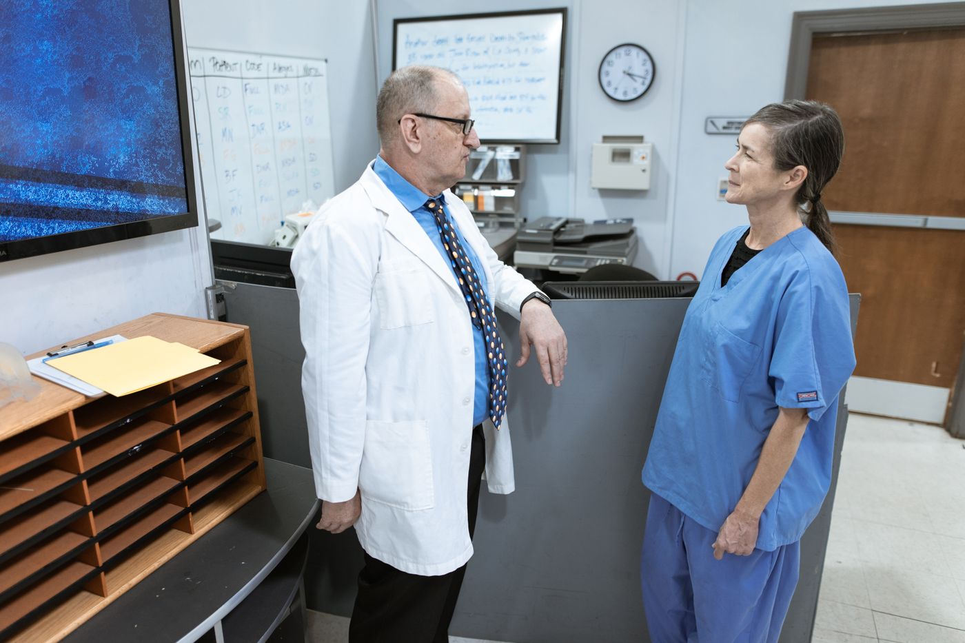 A nurse in blue scrubs talks with a colleague in a white coat beside the nurses' station, with a whiteboard and wall clock behind them