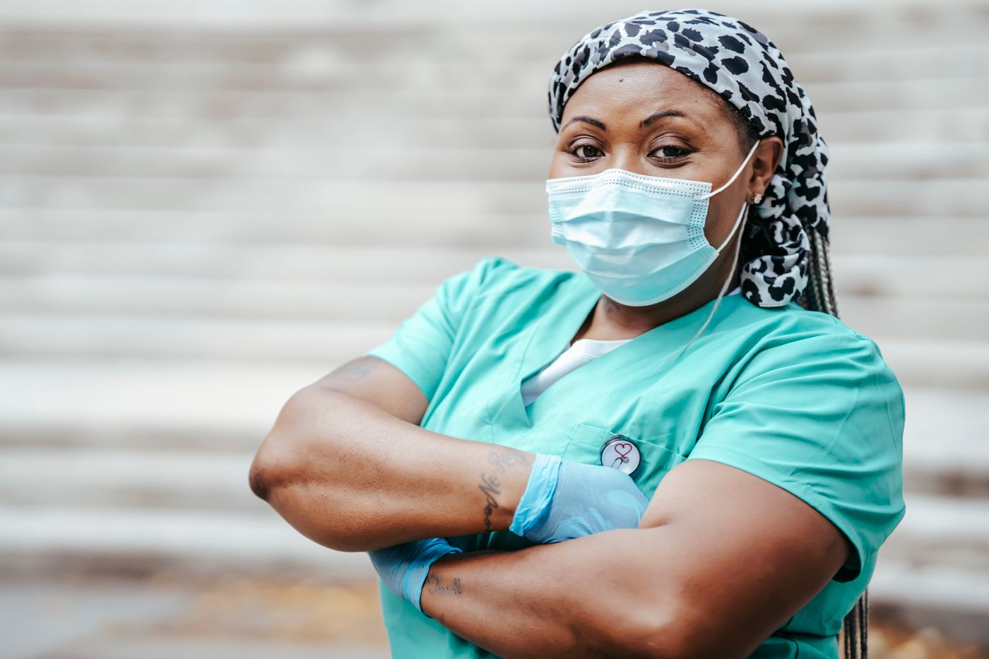 Nurse in teal scrubs, a patterned head wrap and a face mask standing with arms folded and a steady, direct expression