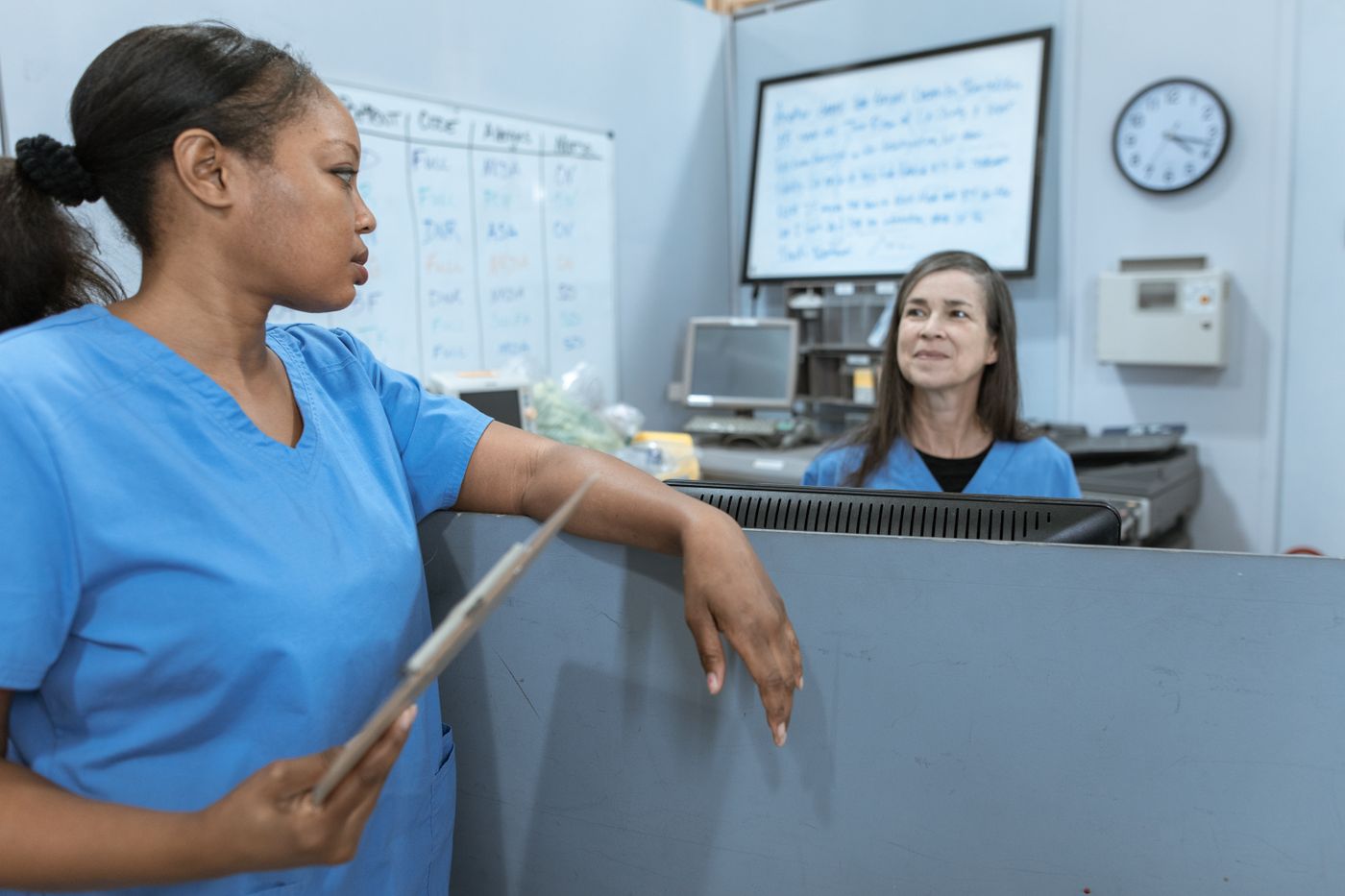 Two nurses in blue scrubs coordinating at a nursing station with an assignment whiteboard and a wall clock behind them