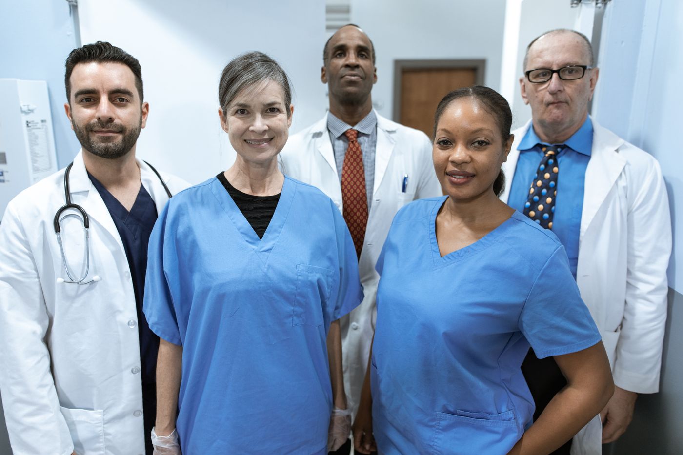 A team of five healthcare professionals in scrubs and white coats standing together in a hospital corridor, looking confidently at the camera