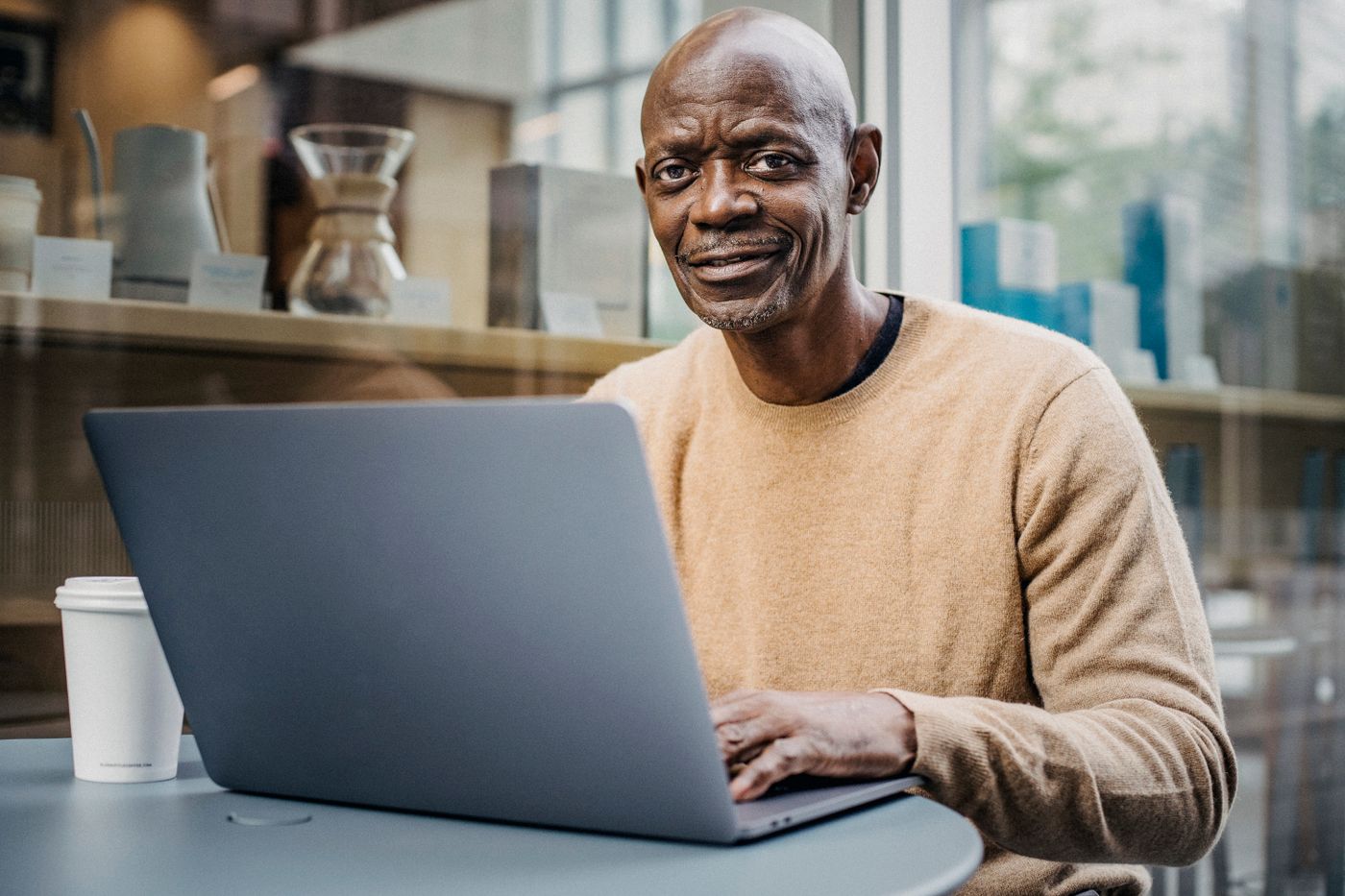 Smiling mature man in a tan sweater researching nursing programs on a laptop at a cafe table