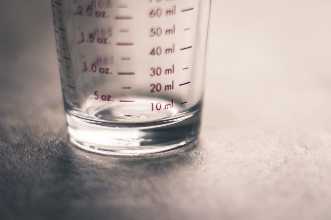 Close-up of a small clear glass measuring beaker on a pale surface, marked with a millilitre scale on one side and a fluid ounce scale on the other