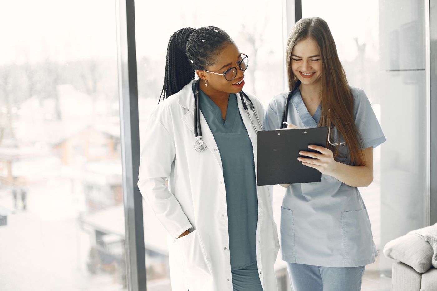An experienced clinician in a white coat and a newer nurse in blue scrubs standing together by a window, reviewing notes on a clipboard and smiling