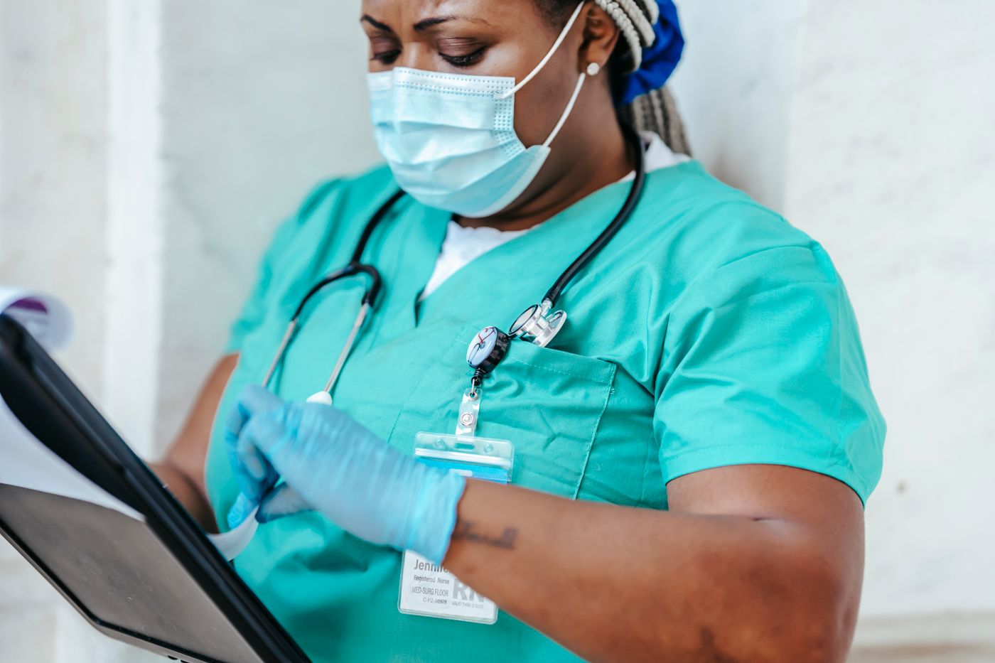 A registered nurse in teal scrubs, a face mask and gloves, with a stethoscope and a hospital ID badge, reads through papers on a clipboard