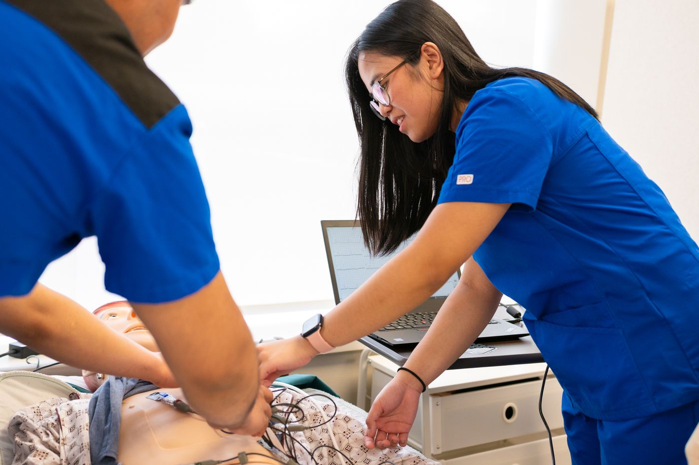 Two nurses in blue scrubs practising chest compressions on a training manikin at a skills station, with a laptop showing a cardiac rhythm trace beside them