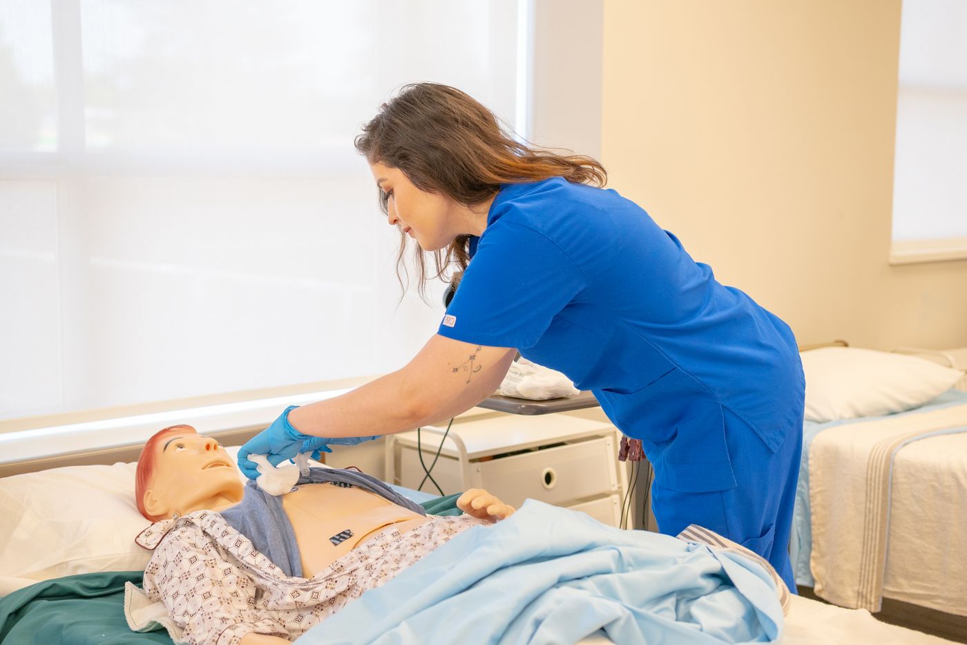 A nurse in blue scrubs and gloves practicing a chest assessment on a patient simulation manikin in a bright hospital skills lab