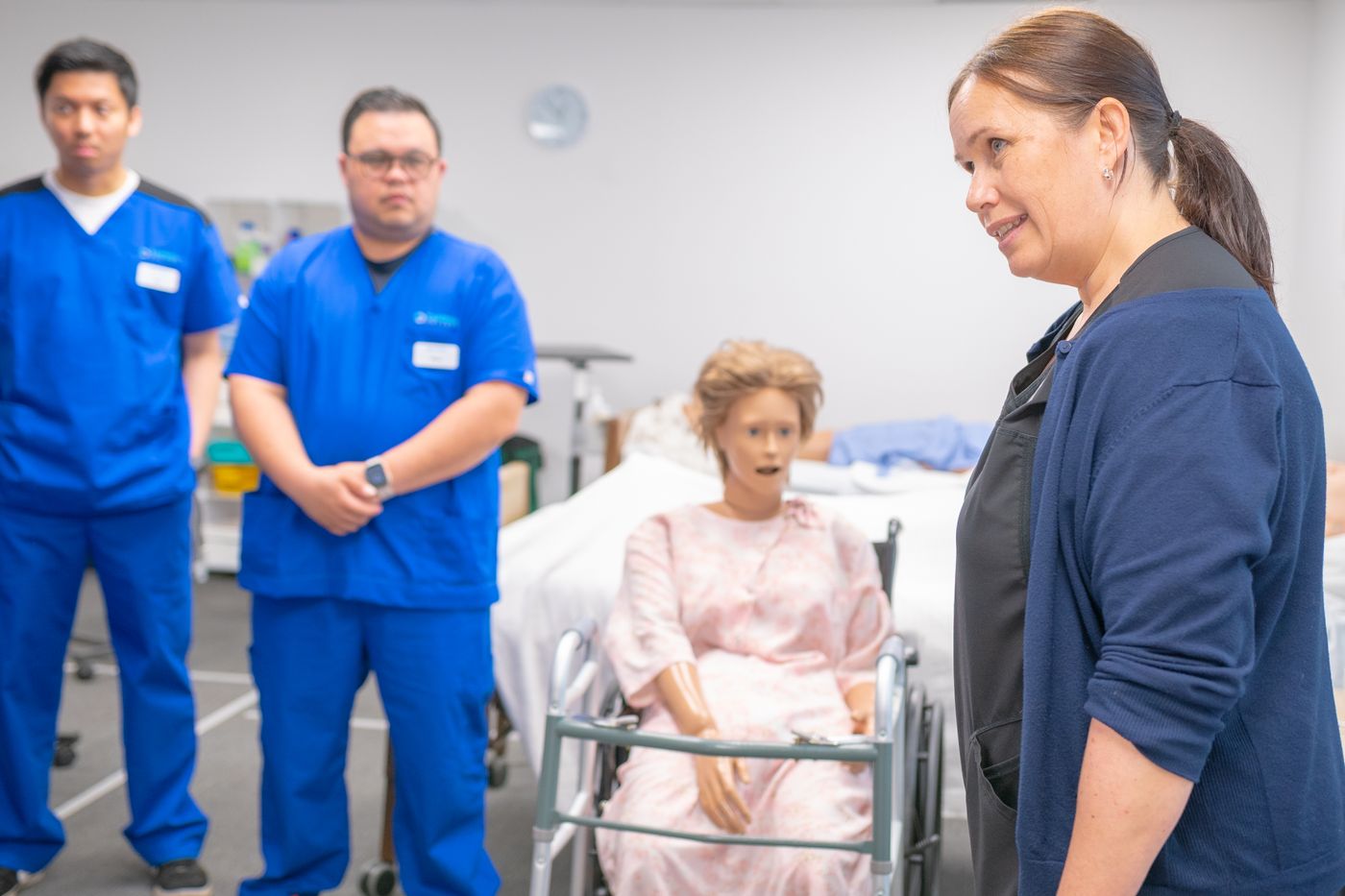 A nurse educator in a navy cardigan teaching two nurses in blue scrubs beside a patient training manikin in a hospital skills lab