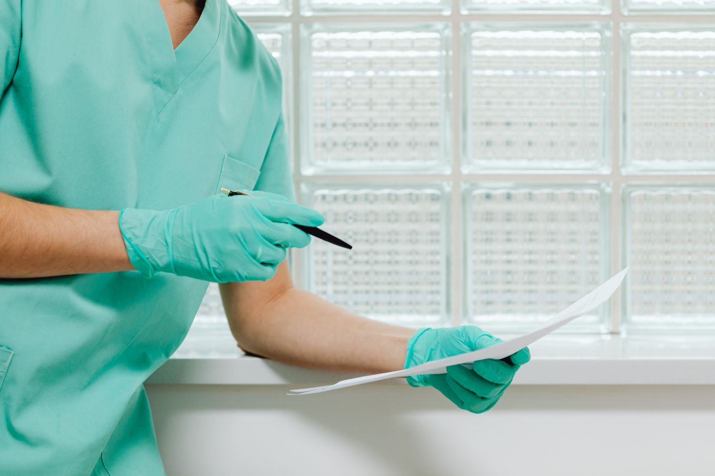 Healthcare worker in green scrubs and gloves holding a printed form and a pen beside a glass-block window