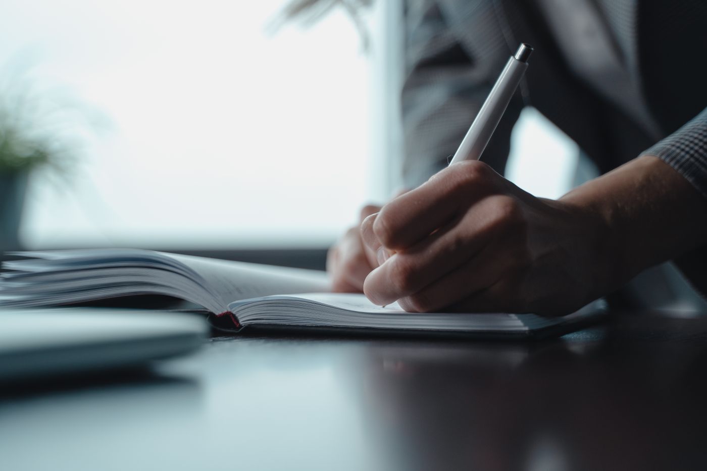 Close-up of a person writing by hand in an open notebook at a desk beside a bright window