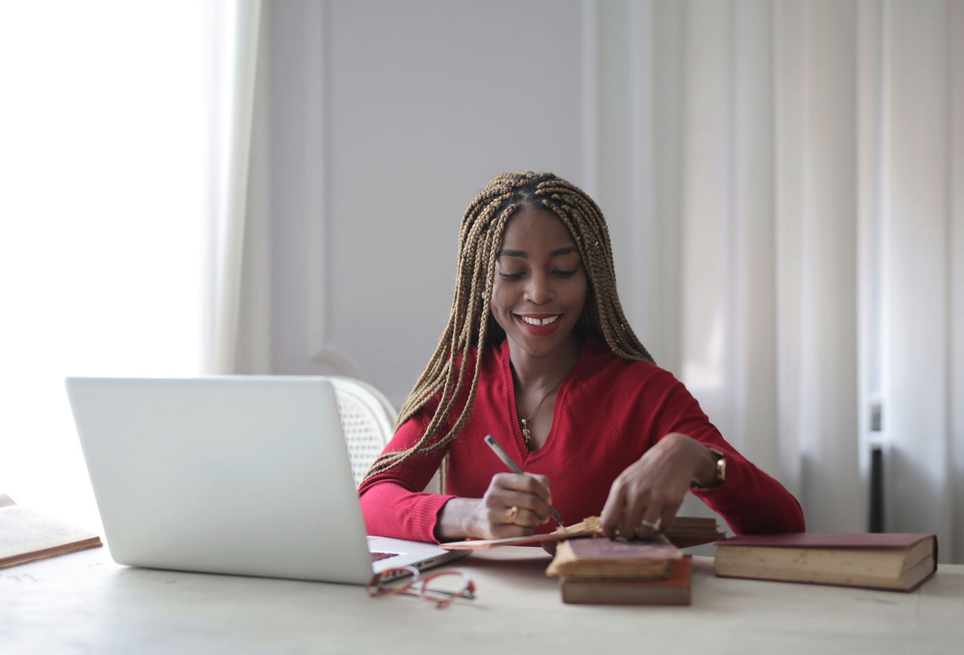 Smiling woman in a red top writing notes beside a laptop and a stack of books while studying at a desk