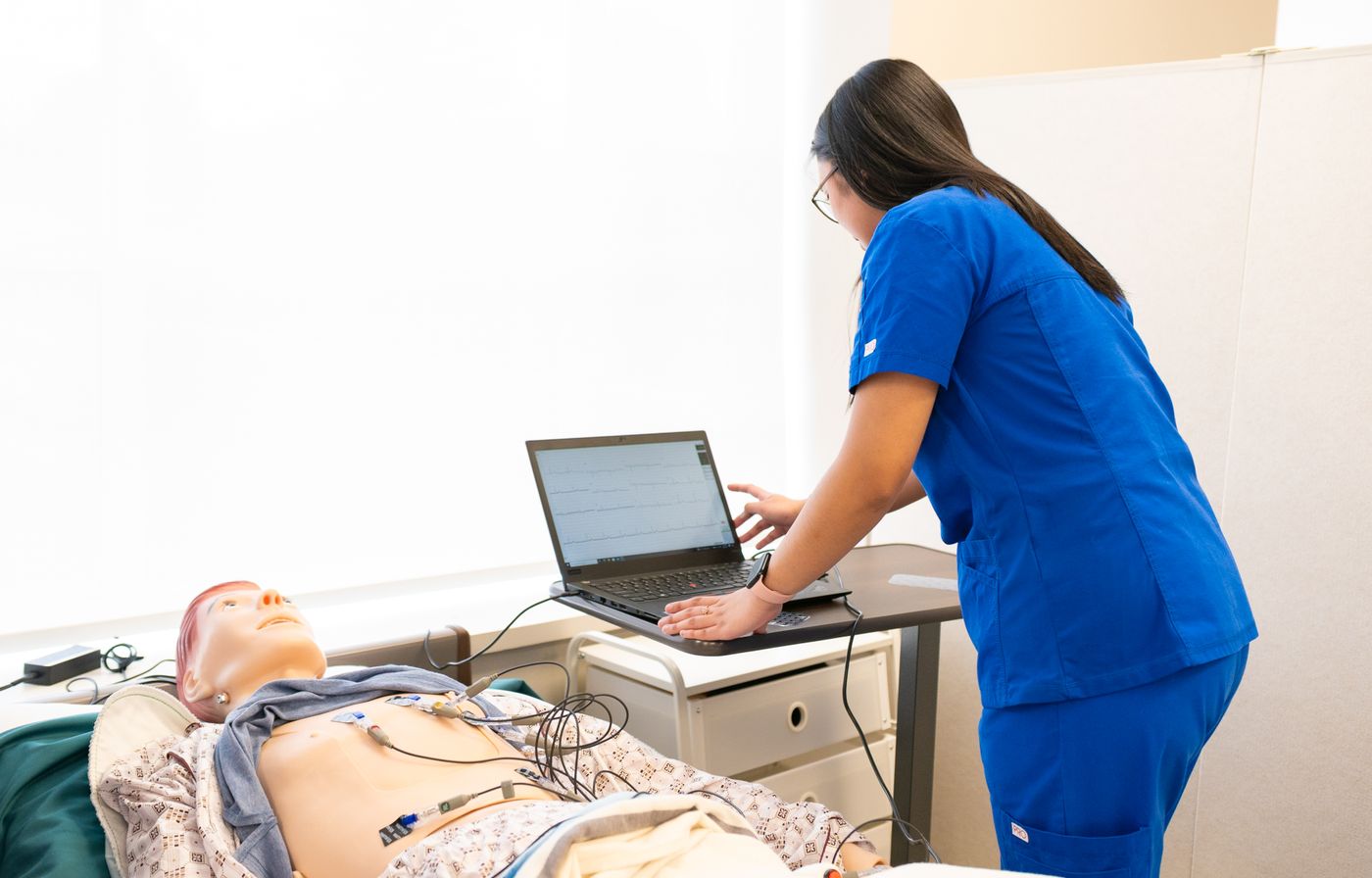 A nursing student in blue scrubs leans over a laptop on a bedside table beside a patient simulation manikin in a bright skills lab